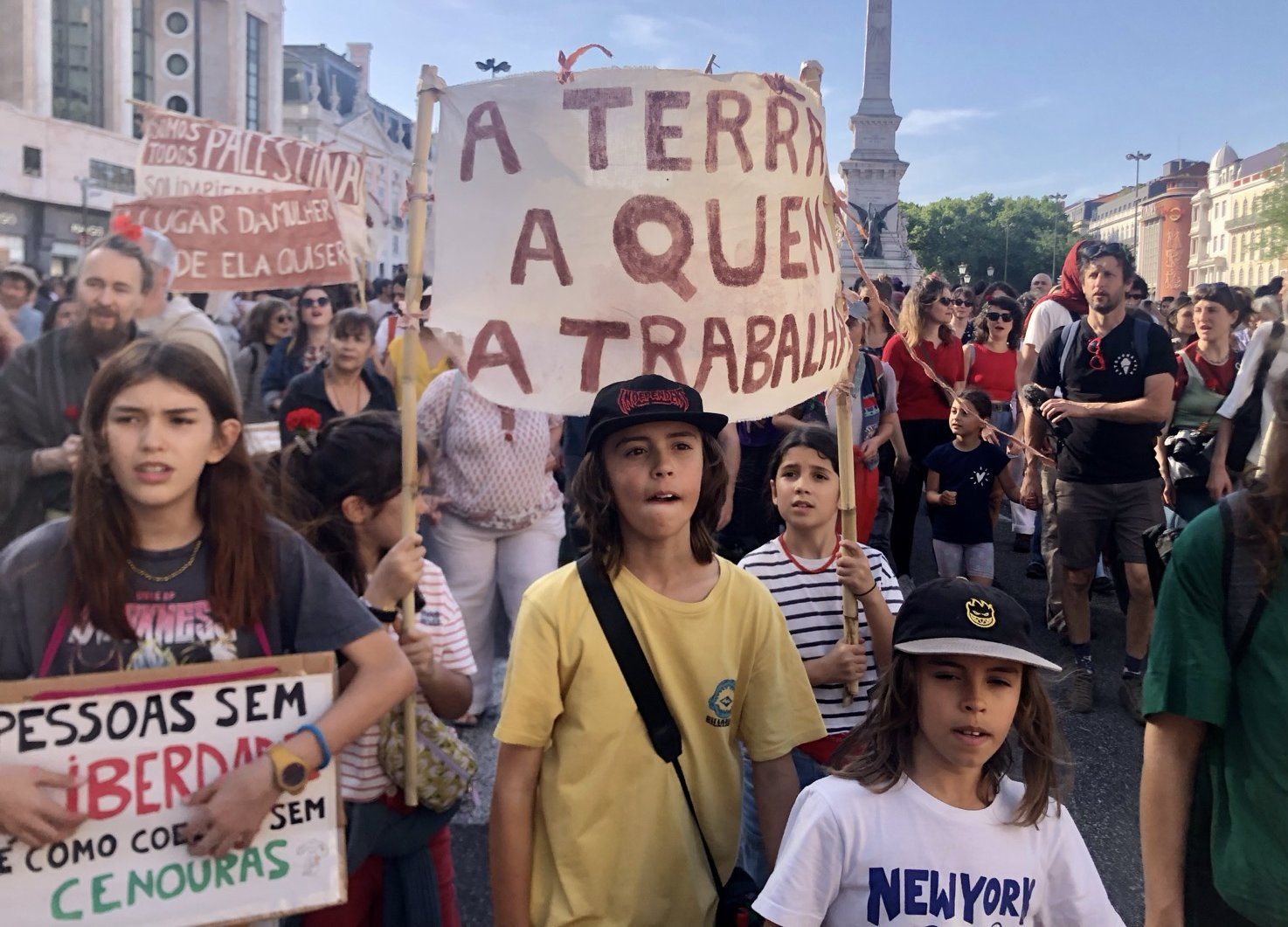Várias crianças com vários cartazes. No plano principal um cartaz que lê "A Terra A Quem A Trabalha"