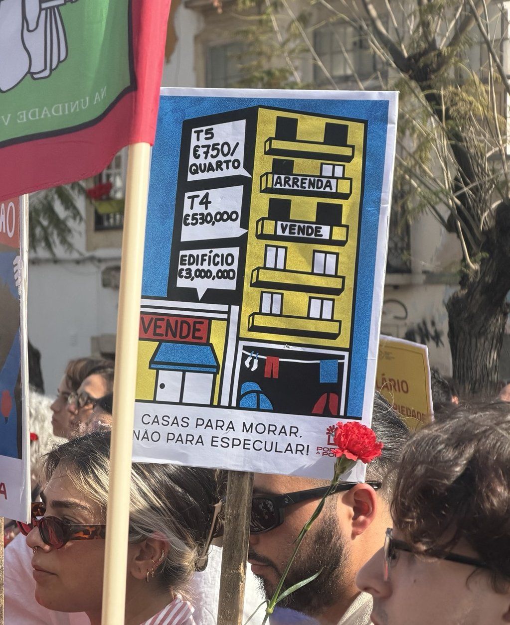 Dois jovens seguram num cartaz, atrás de um cravo vermelho, com uma ilustração de um prédio com preços exurbitantes mas reais