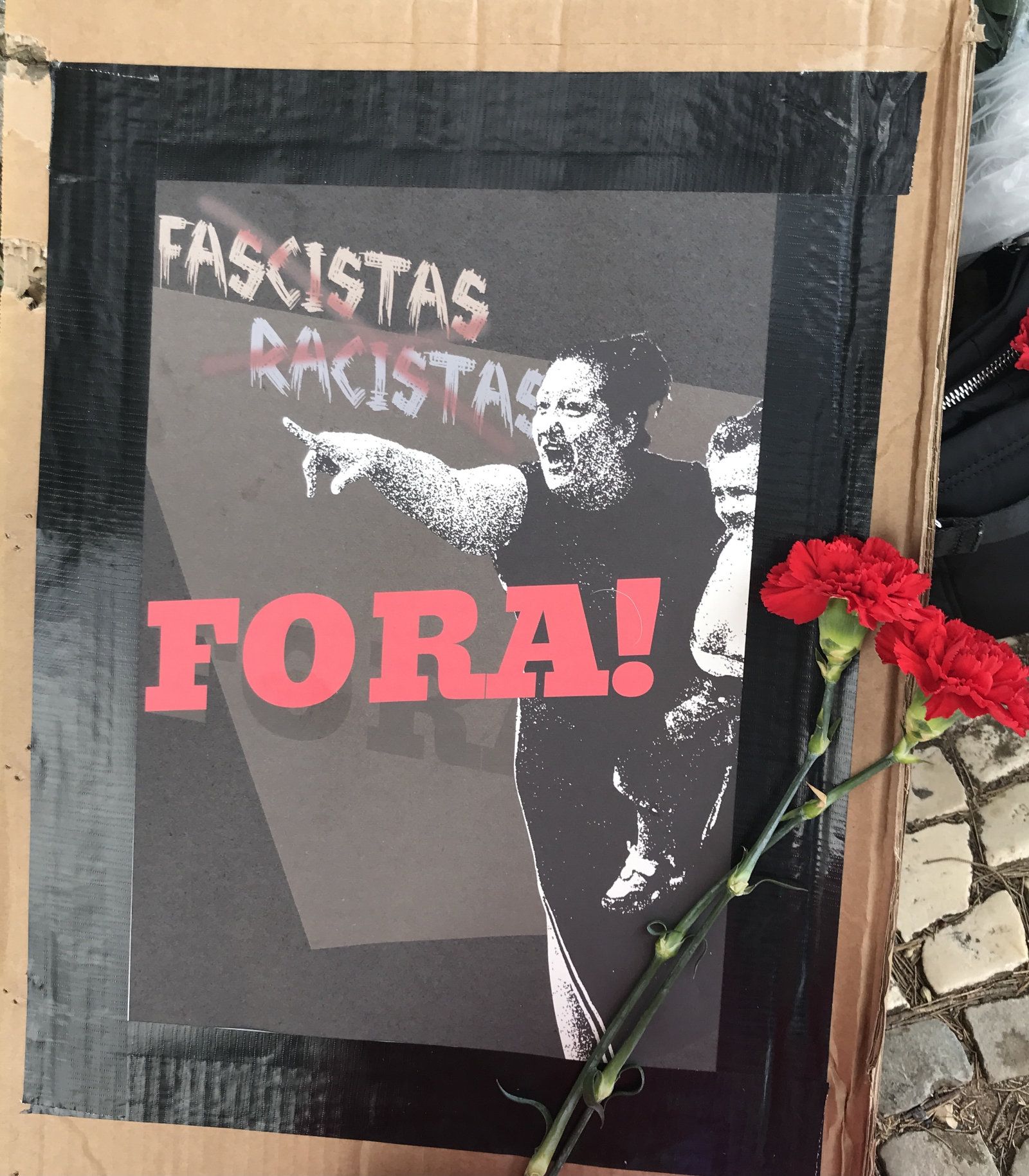 Um cartaz feito com colagens tem uma senhora a apontar para a rua e a frase "Fascistas e Racistas Fora!". Encostado ao cartaz estão dois cravos vermelhos