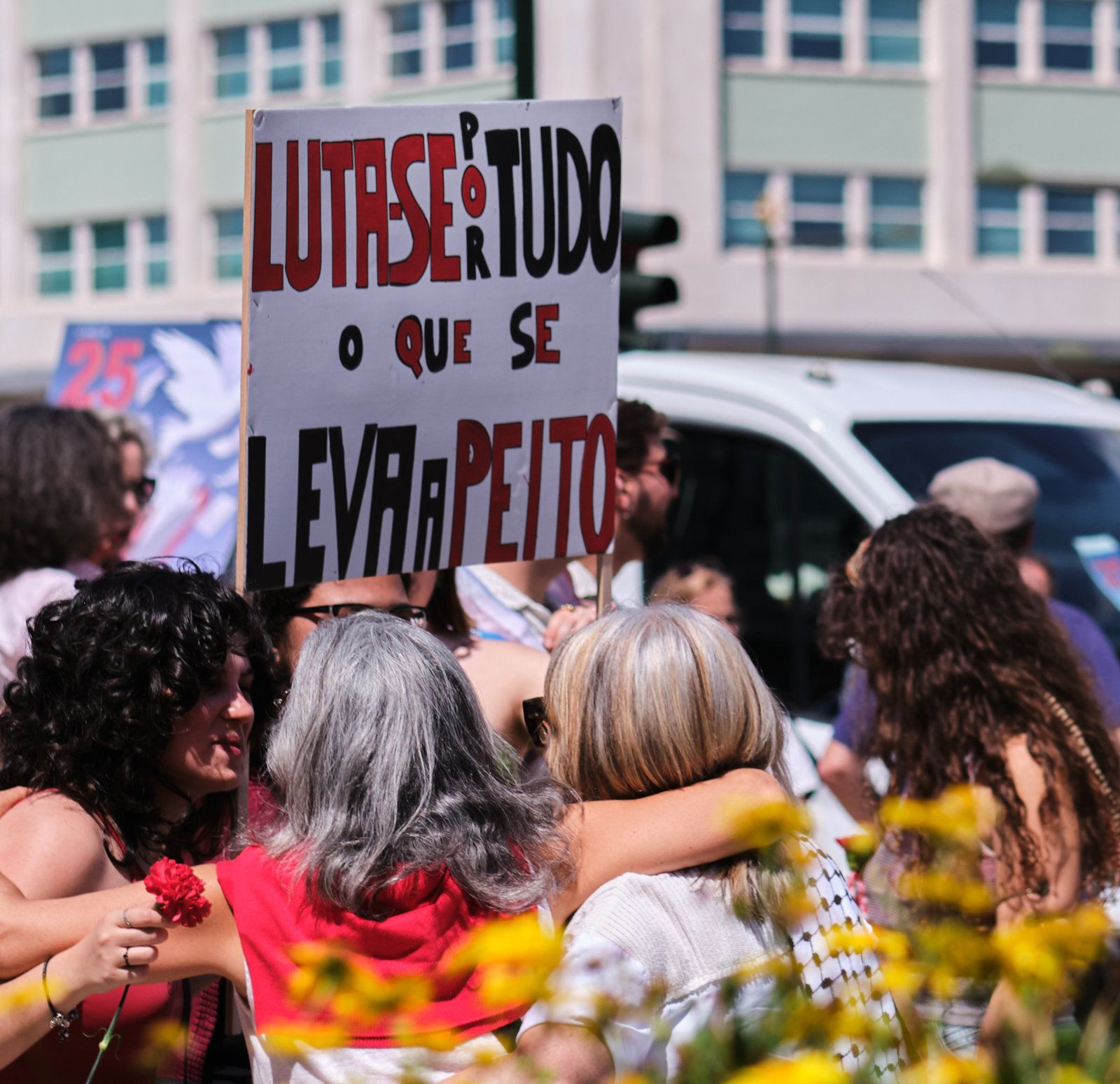 Três mulheres de diferentes idades abraçam-se enquanto seguram um cartaz que diz "Luta-se Por Tudo Que Se Leva a Peito"