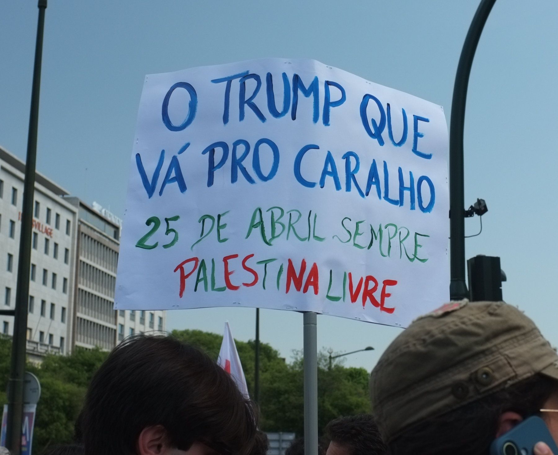 Uma cartaz na multidão lê "O trump que vá pro caralho, 25 de abril sempre, palestina livre"