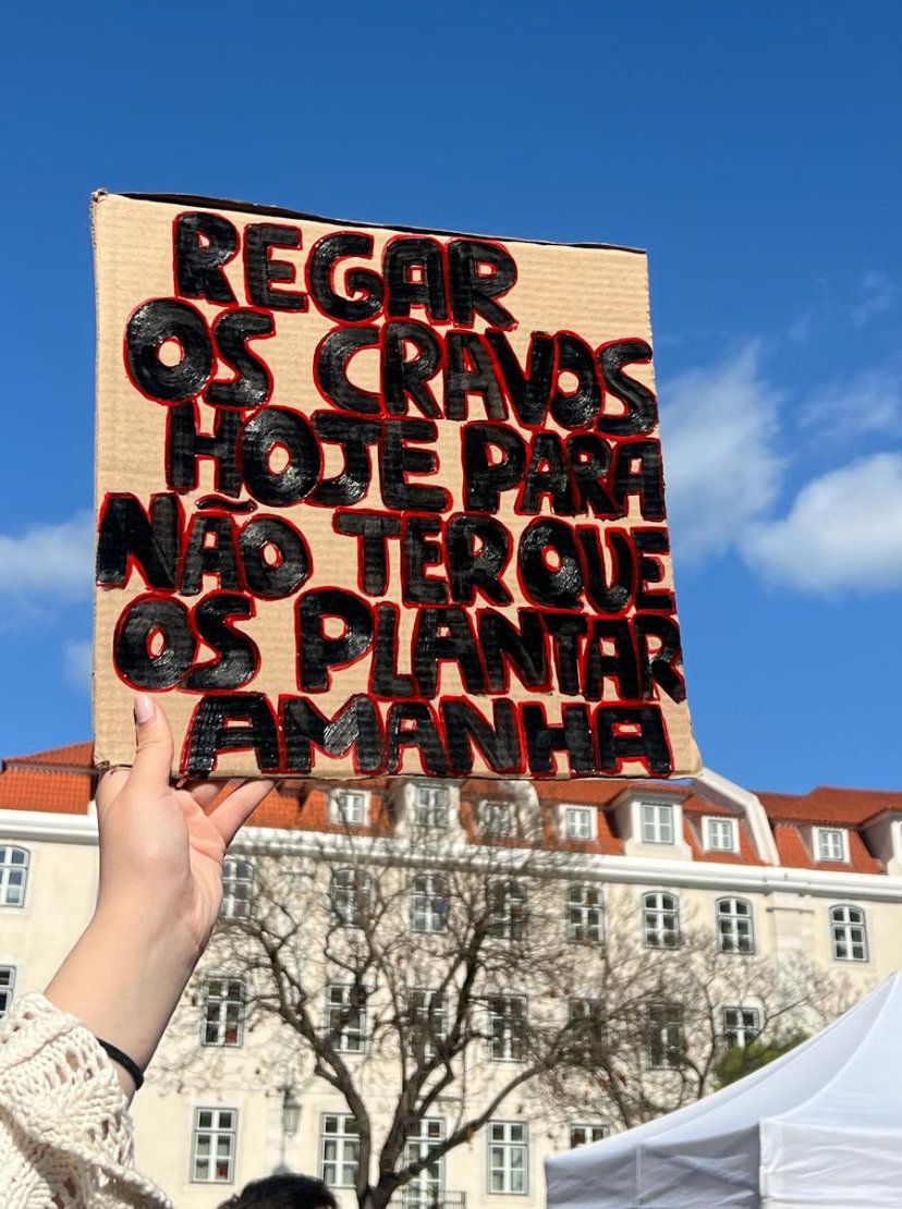 Uma mão segura um cartaz que diz "Regar os cravos hoje para não ter que os plantar amanhã"