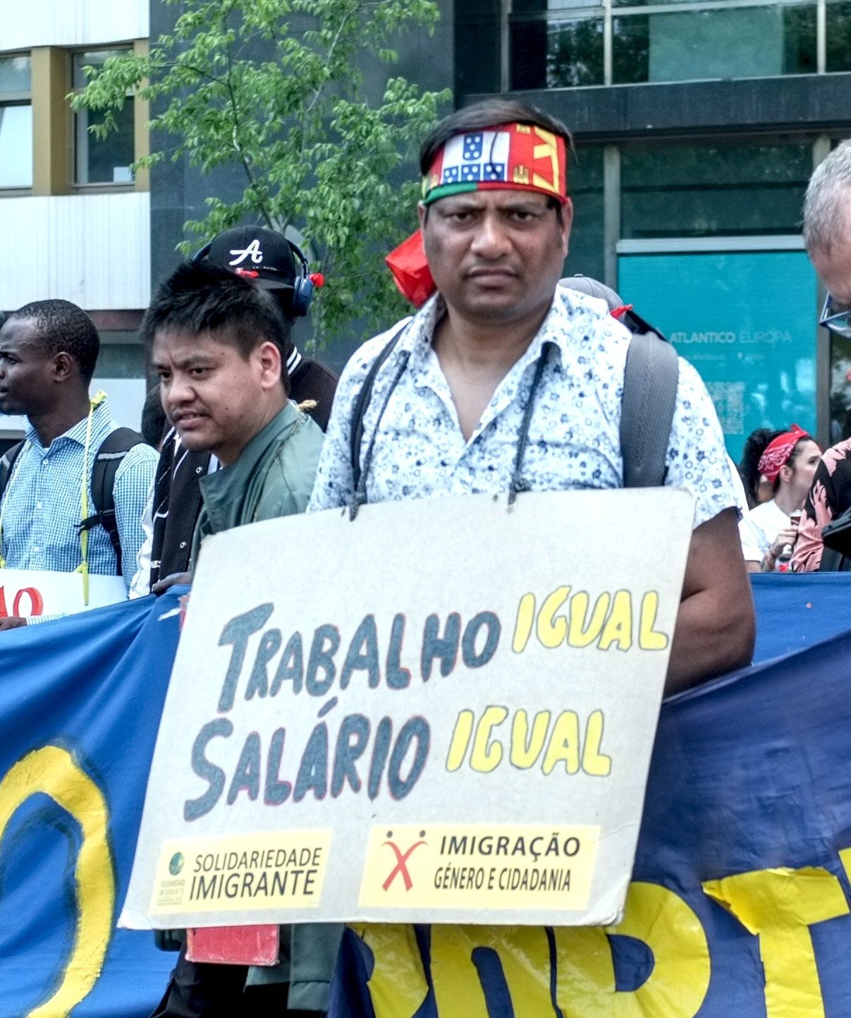 Um senhor não português de origem segura um cartaz que diz "Trabalho igual, salário igual. Solidariedade imigrante. Imigração género e cidadania". Atrás, outras pessoas racializadas.