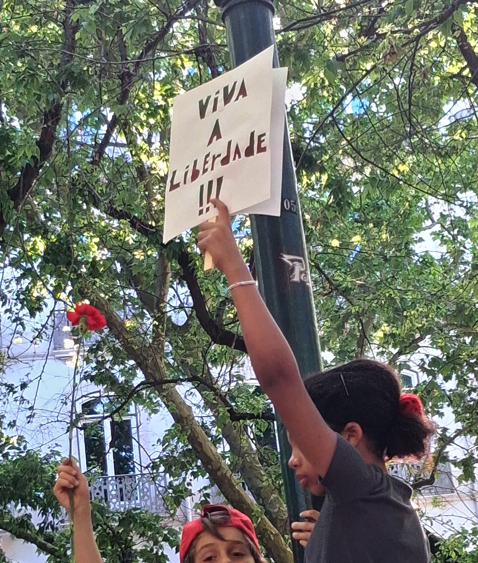 Duas crianças estão na foto, uma segura um cravo vermelho bem alto, e a outra um cartaz no qual se lê "Viva a Liberdade!"